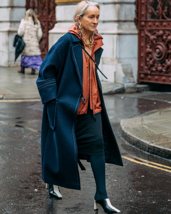 Femme de plus de 60 ans avec manteau long sombre et veste zippée, portant des chaussures à talons qui allongent la silhouette