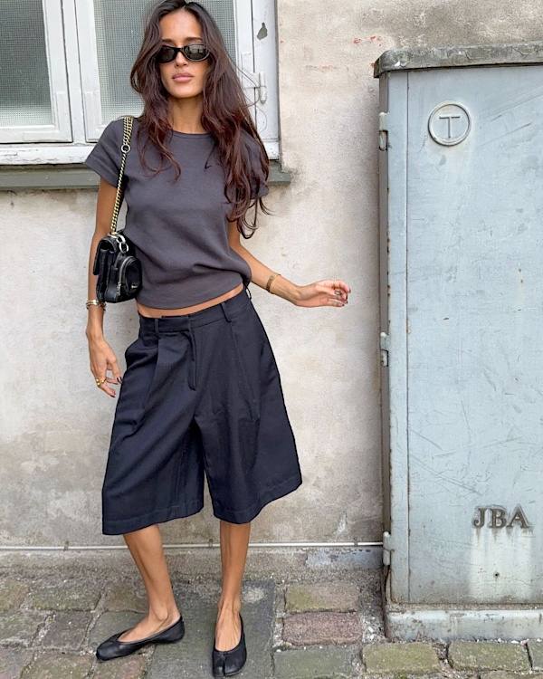 Femme portant un bermuda noir ample avec un tee-shirt gris, des ballerines noires et un petit sac à chaîne