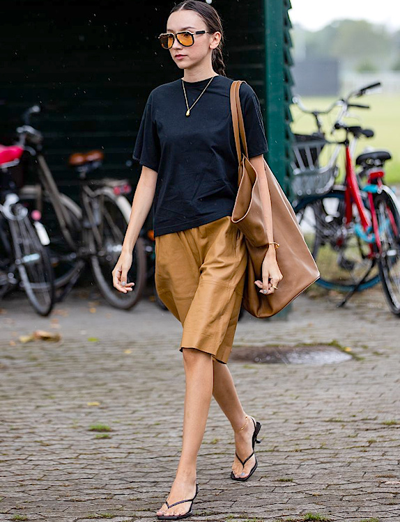 Femme portant un bermuda camel souple avec un tee-shirt noir, un grand sac en cuir et des tongs fines
